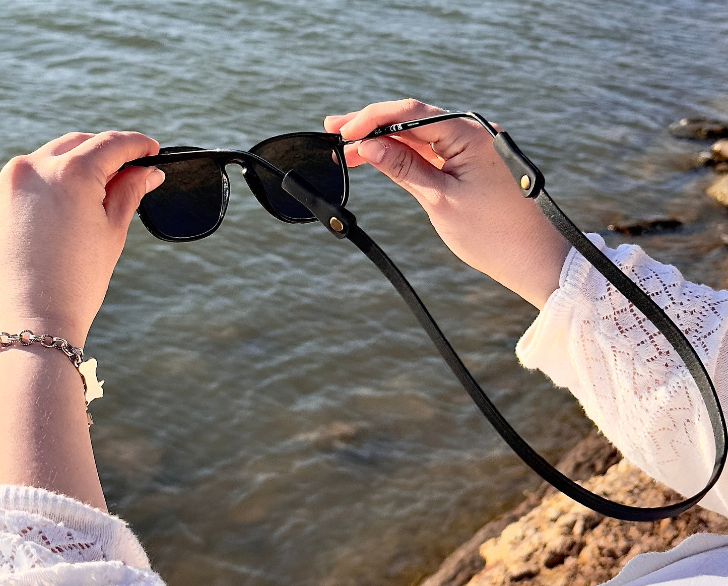 Person holding sunglasses with a scenic background of water and rocks.