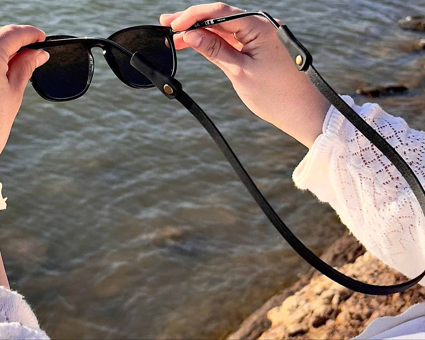 Person holding sunglasses with a scenic background of water and rocks.
