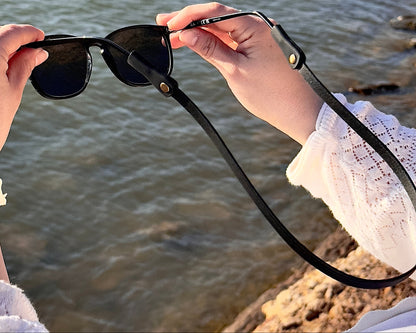 Person holding sunglasses with a scenic background of water and rocks.